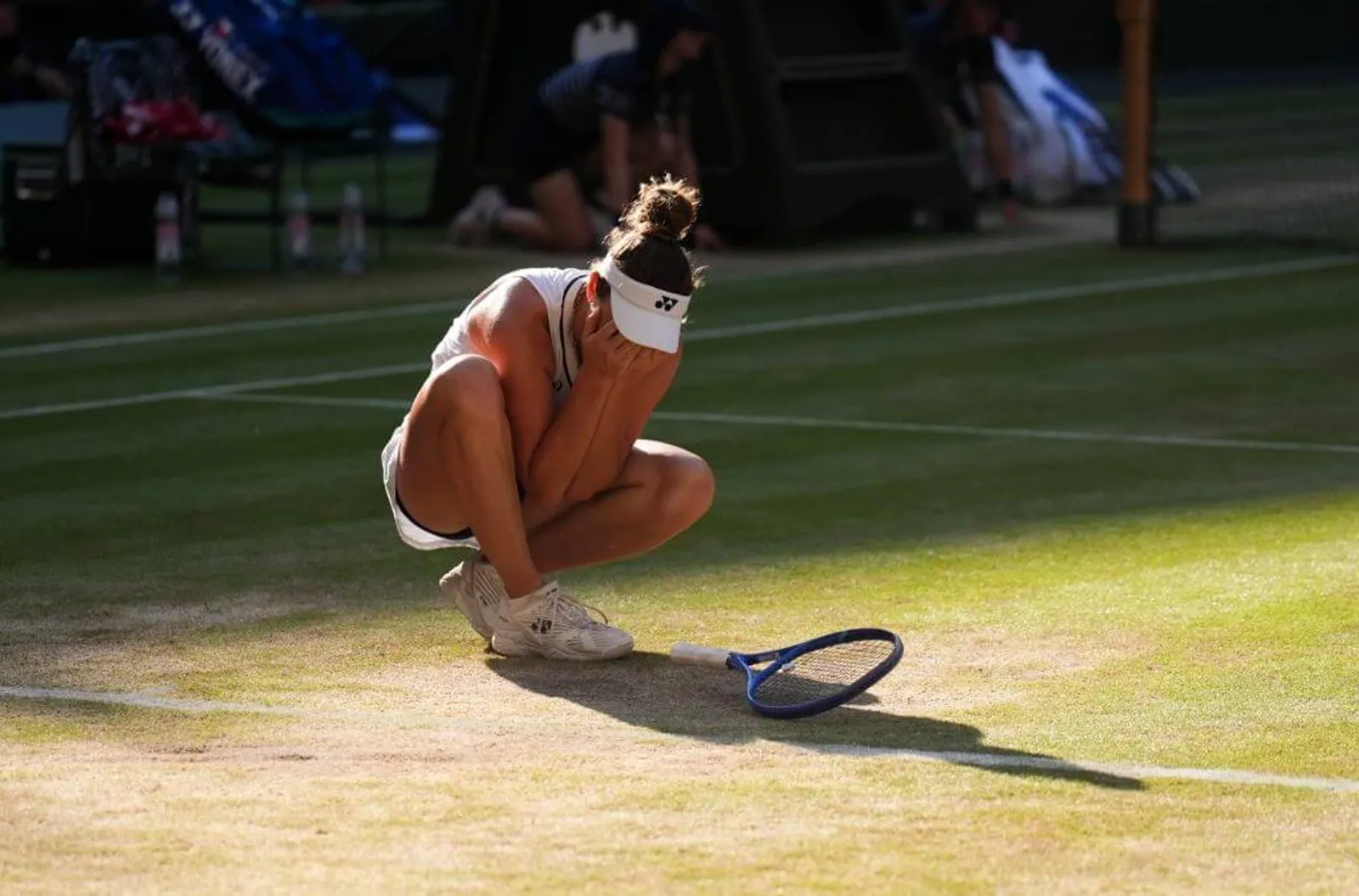 Linda Nosková sits on her haunches on a grass court with her hands over her face in joy.